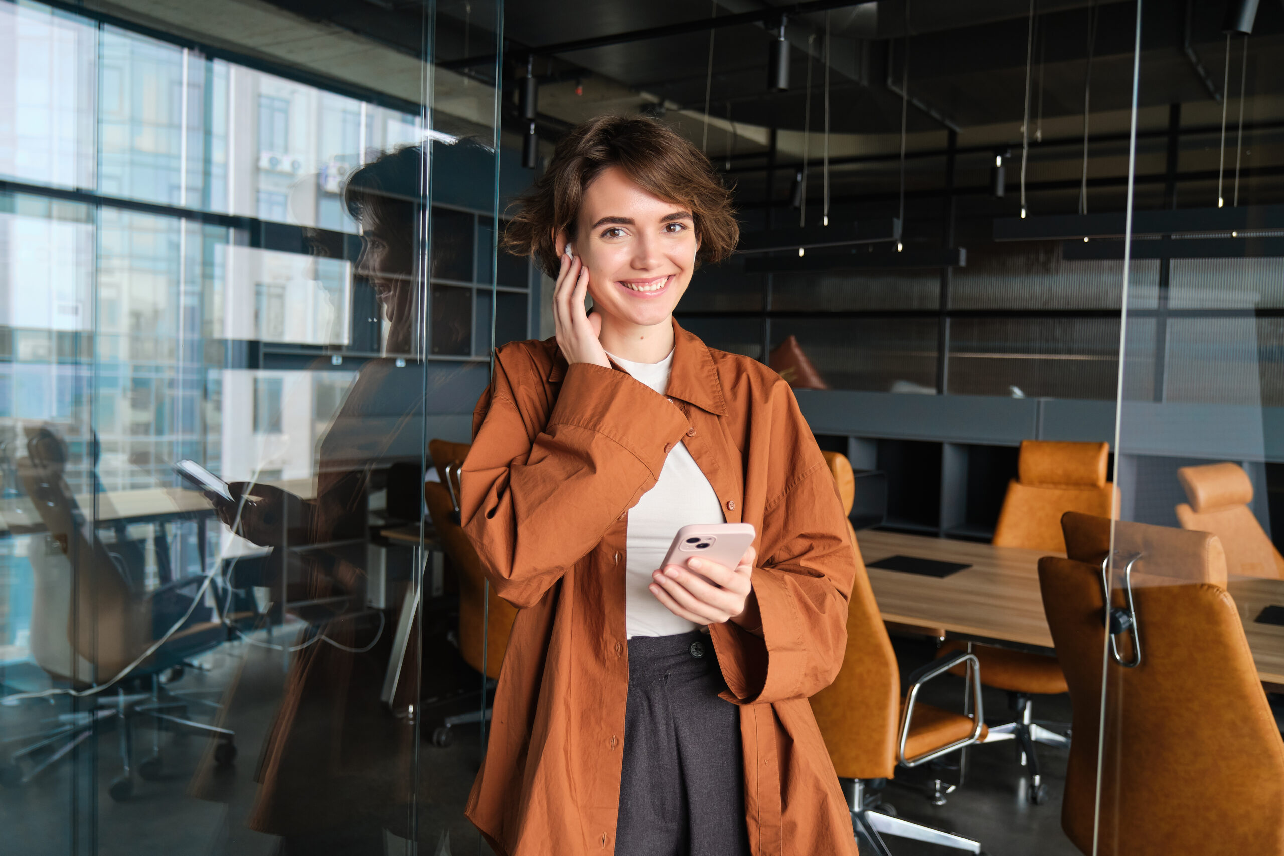 Image of woman calls someone on phone, stands with smartphone and wireless earphones, poses in office and smiling